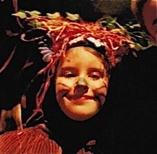 A child in woodlander costume, including a headdress decorated with leaves, flowers and colourful straw.