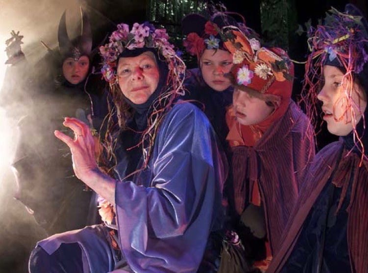 Marna and the Woodlanders, wearing floral headdresses, with the black-clad figure of Otis Claw lurking in the shadows.