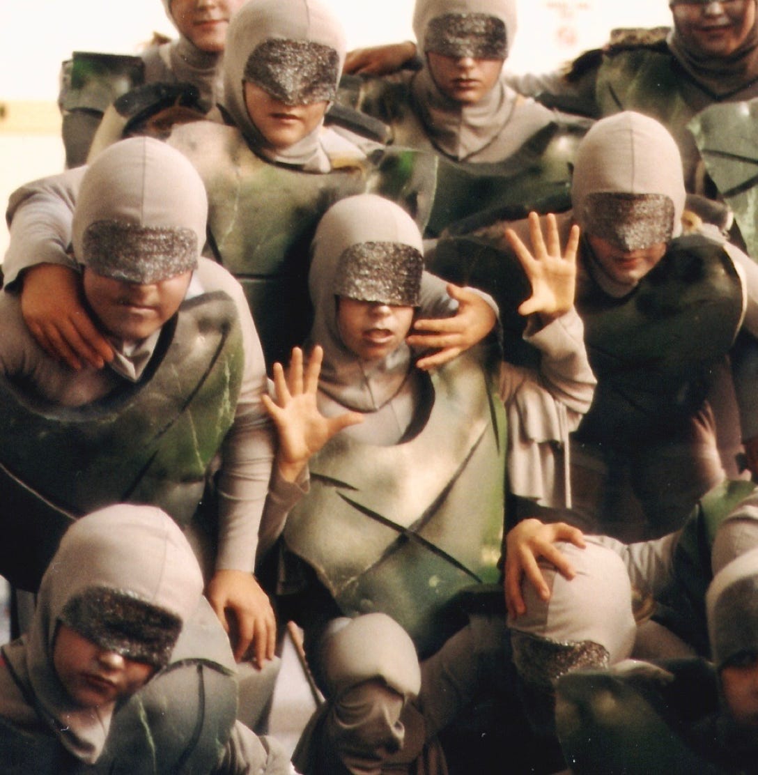 A group of children in grey costumes with padded chestplates and masked eyes.